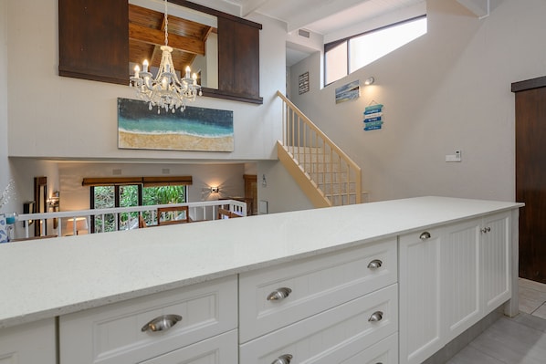 A spacious kitchen with a large island, wooden ceiling beams, a chandelier, and a staircase leading to an upper level.