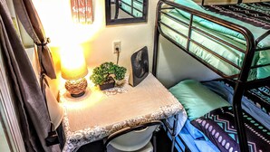 A dormitory room with bunk beds, a bedside table with a lamp, a small potted plant, and a wall-mounted mirror.