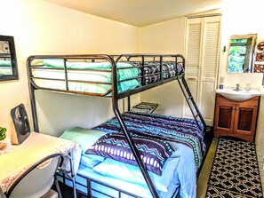 A bunk bed with green and blue bedding, a small desk with a chair, a mirror, and a sink in a compact room. - River View Rainforest Tiny Cabin with Kitchenette (No Cleaning Fee) (Pepeekeo)