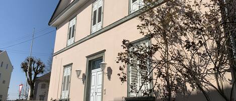 A two-story building with a white door and shutters, a staircase with a metal railing, and a tree in front.