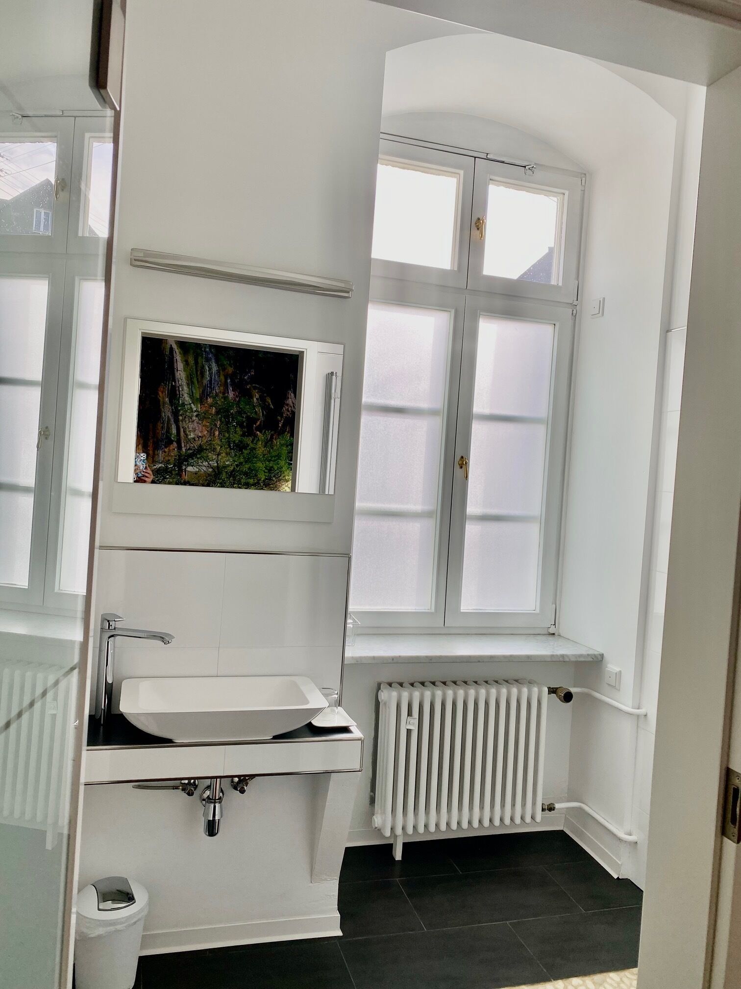 A bathroom with a white sink, a window with frosted glass, a radiator, and a trash bin.