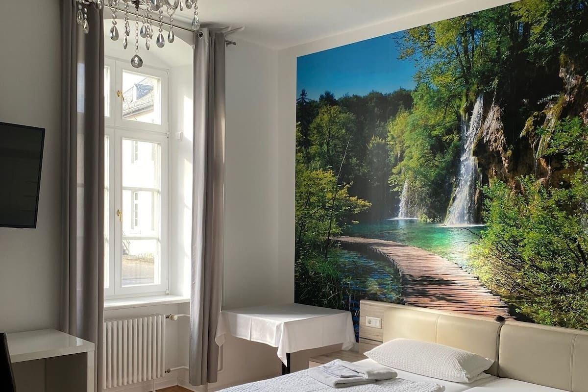 A bedroom with a large bed, a desk, a chair, a chandelier, and a wall mural of a waterfall.