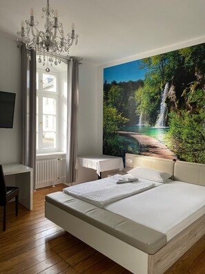 A bedroom with a large bed, a desk, a chair, a chandelier, and a wall mural of a waterfall. - Frankfurter Hof 1834 (Wallmerod)