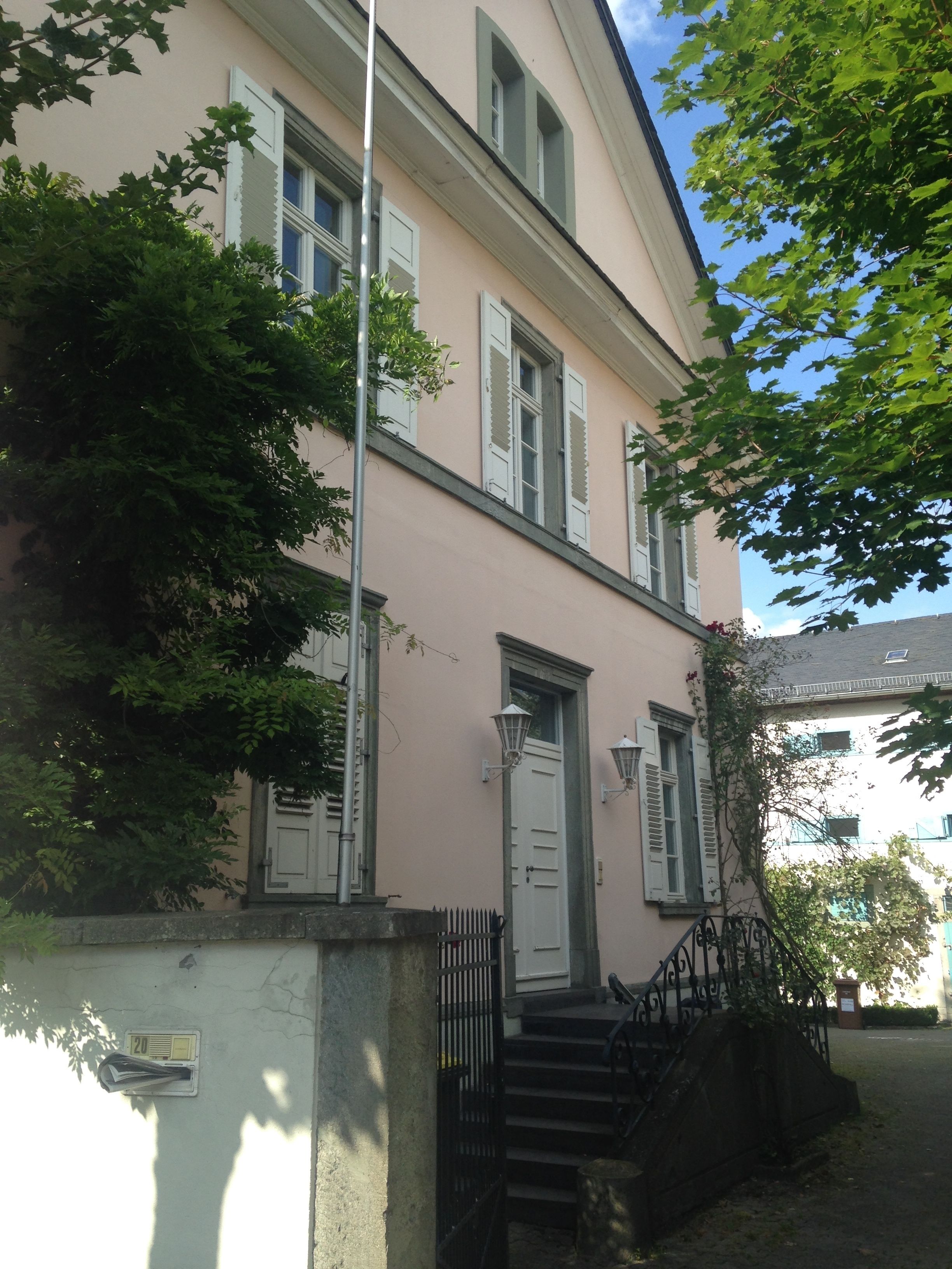 Ein zweistöckiges Gebäude mit einer rosa Fassade, weißen Fensterläden und einem kleinen Eingangsbereich mit Stufen.
