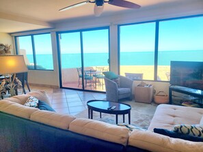 A living room with a large window overlooking the sea, a sofa, a television, and a ceiling fan.