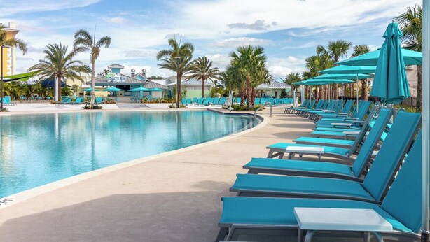 A poolside area with lounge chairs and umbrellas, palm trees, and a building in the background.