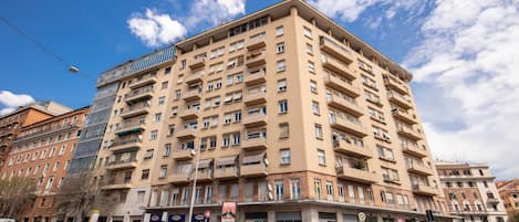 Un immeuble à plusieurs étages doté d’une façade moderne en verre, de nombreux balcons et d’un espace commercial au rez-de-chaussée, comprenant des boutiques et des voitures garées.