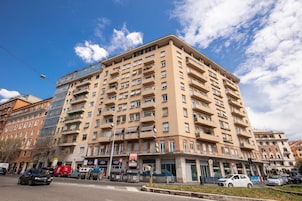 Un edificio a più piani con una moderna facciata in vetro, diversi balconi e una zona commerciale a livello strada con negozi e auto parcheggiate.