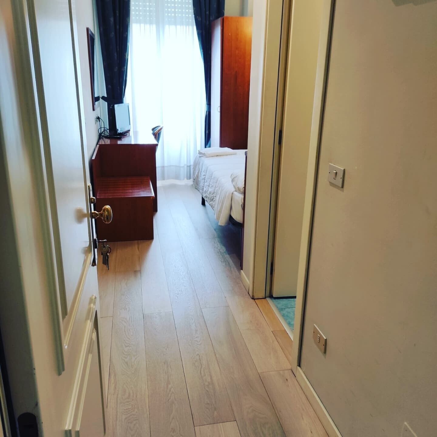 A hotel room with a wooden floor, a bed, a desk with a chair, and a TV.