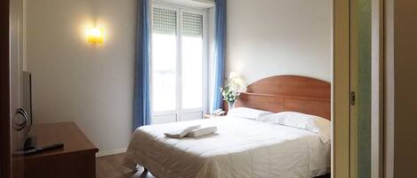 Habitación de hotel con piso de madera, cama con sábanas blancas, cabecera de madera, mesita de noche con lámpara y ventana con cortinas azules.