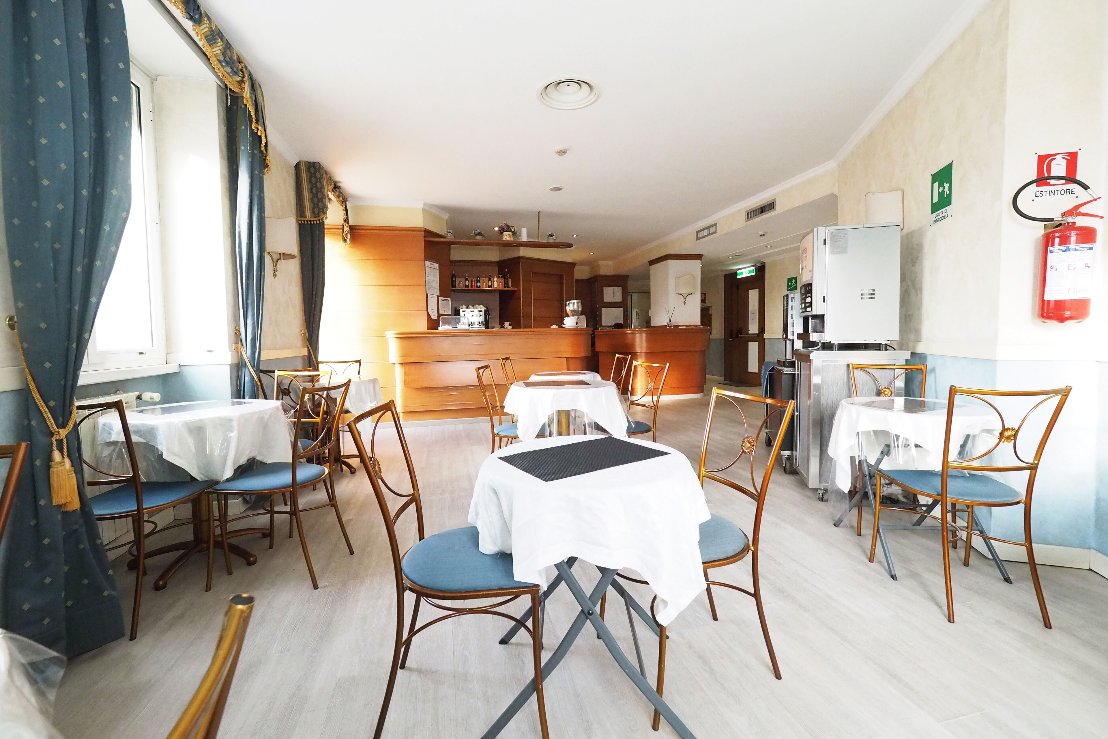 A hotel dining area with tables and chairs, a kitchen in the background, and a fire extinguisher on the wall.