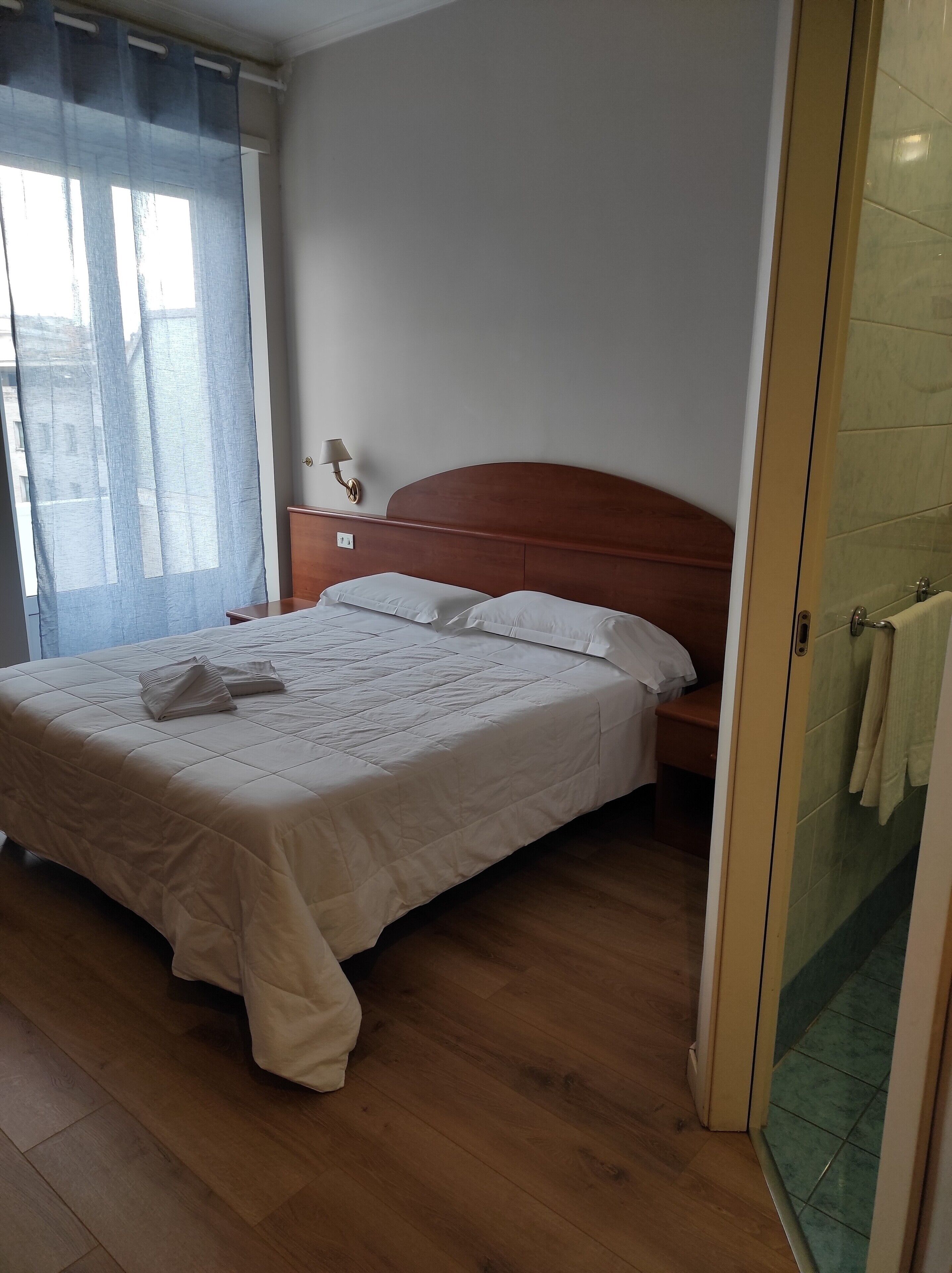 A neatly made bed with a wooden headboard, a bedside table with a lamp, and a view of the outside through a window with sheer curtains.