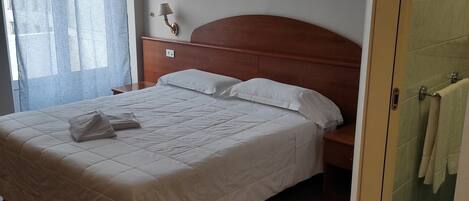 A neatly made bed with a wooden headboard, a bedside table with a lamp, and a view of the outside through a window with sheer curtains.