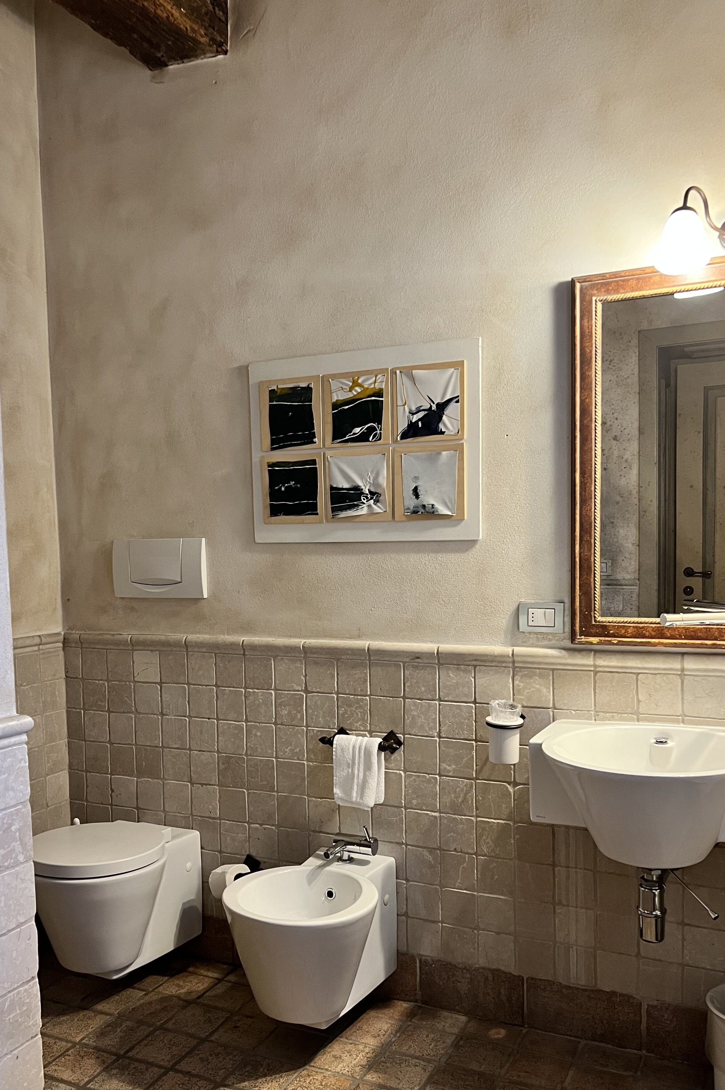 Une salle de bain comprenant des toilettes, un bidet, un lavabo et un miroir. Des tableaux encadrés ornent le mur, et un luminaire est placé au-dessus du miroir.