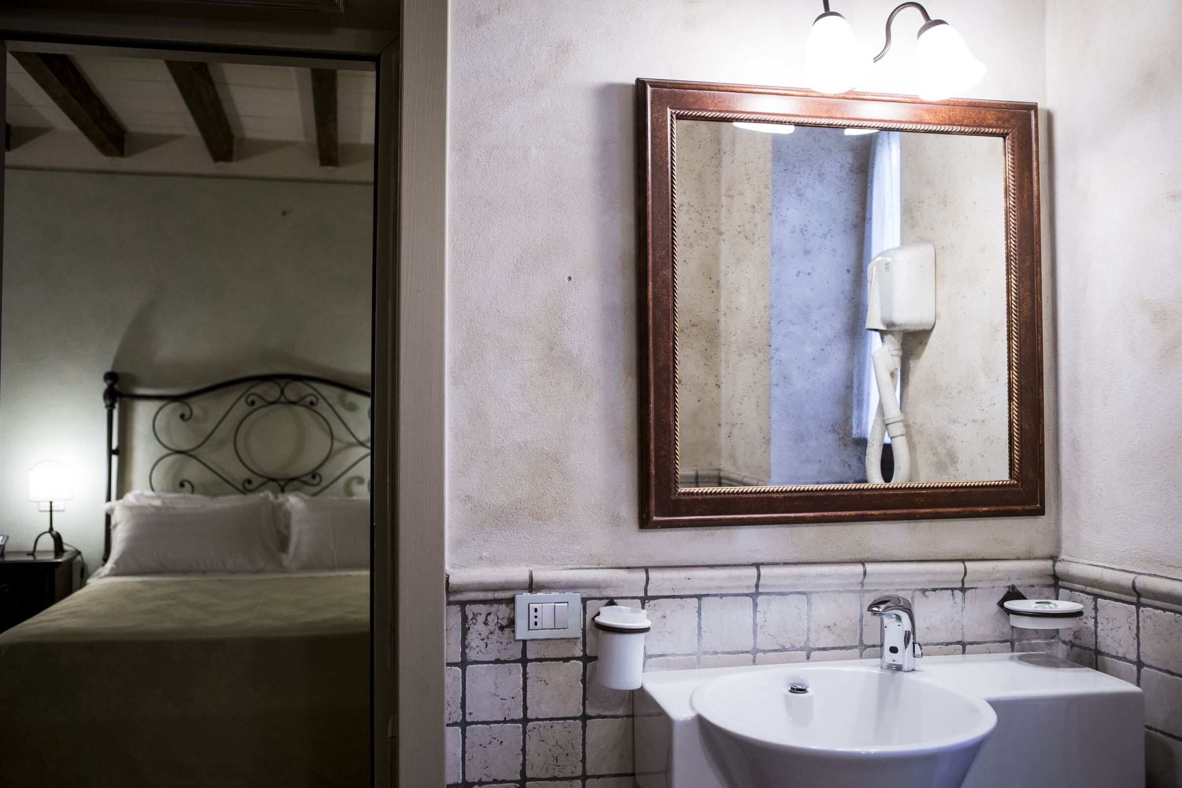 Un baño de hotel con un espejo grande, dos lavamanos y una cama visible en la habitación contigua.