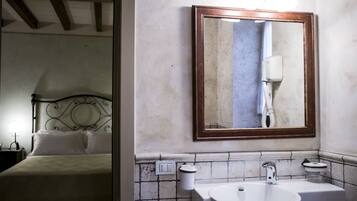 Un baño de hotel con un espejo grande, dos lavamanos y una cama visible en la habitación contigua.