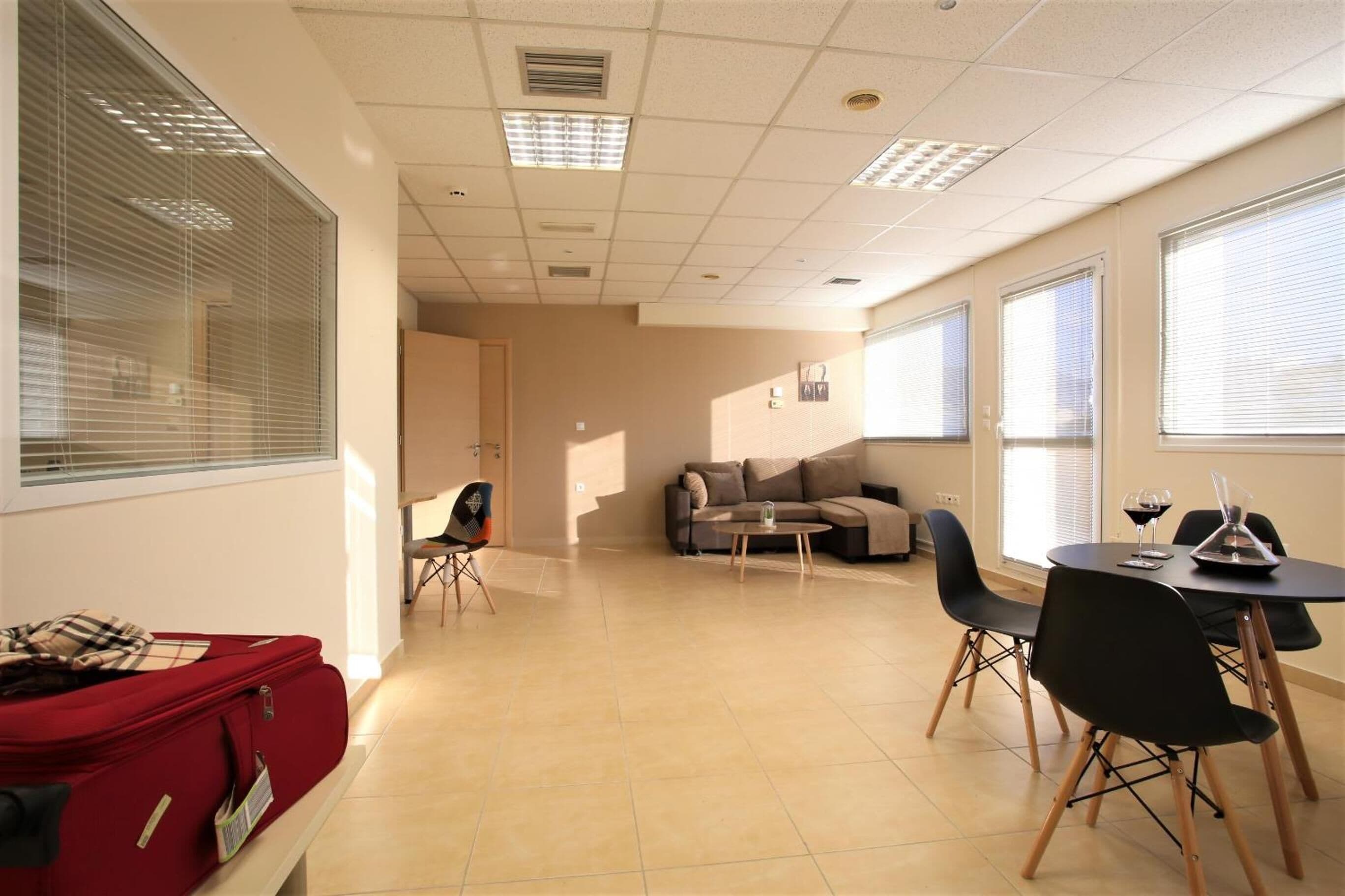 Une pièce spacieuse avec une grande fenêtre, un canapé, une table à manger avec des chaises et une valise rouge posée sur un banc.