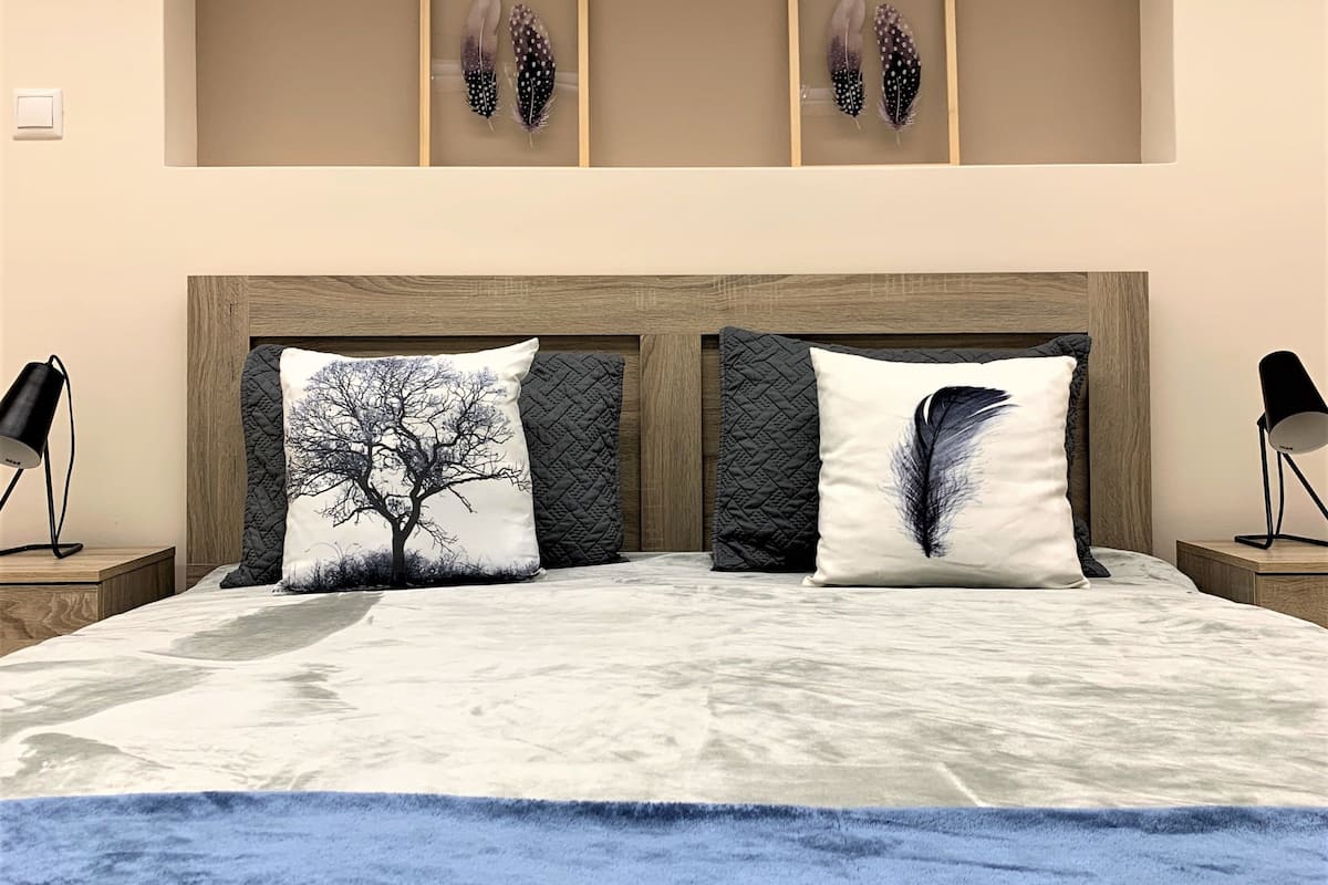 A bedroom with a large bed, two pillows with tree designs, a blue throw blanket, and two framed feather artworks on the wall.
