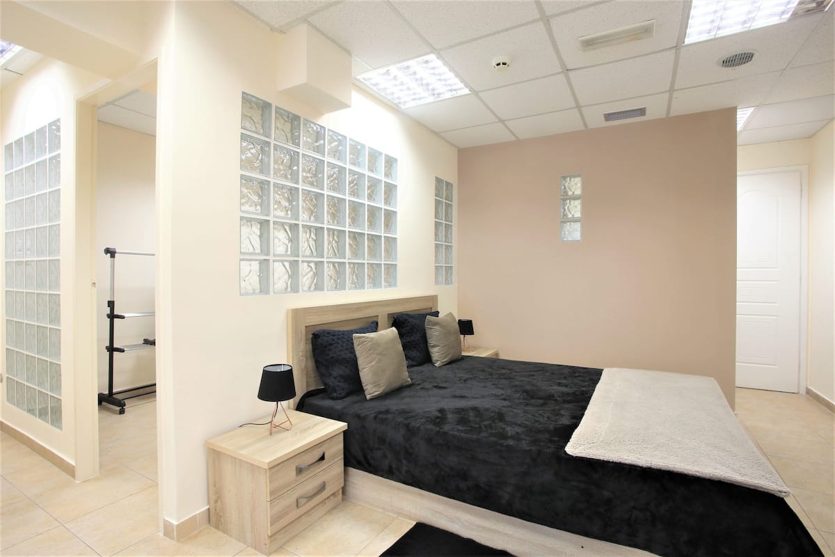 A bedroom with a large bed, a bedside table with a lamp, a window with decorative glass, and a door.