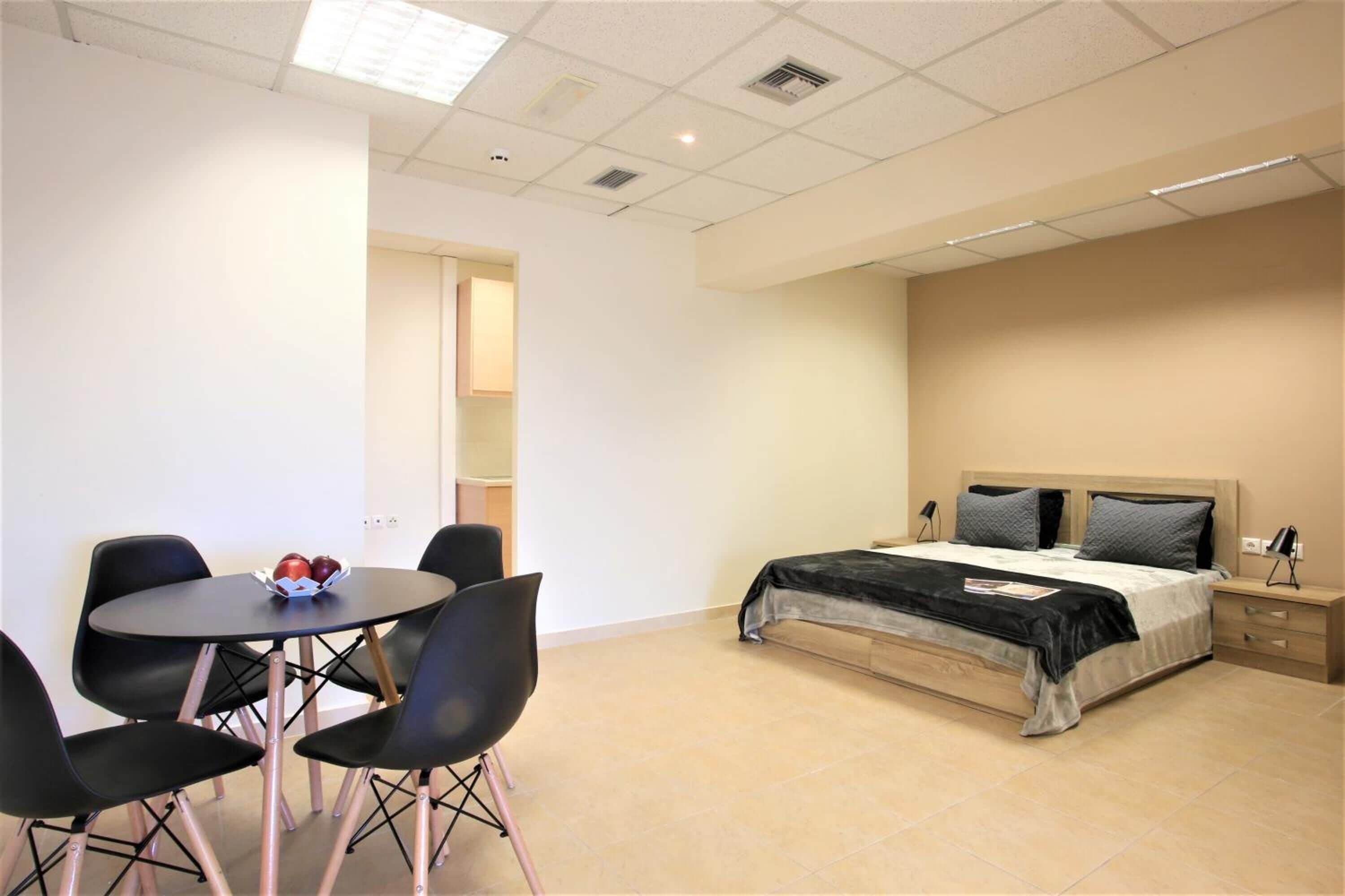 A bedroom with a bed, bedside tables, a nightstand, a desk with chairs, and a small table with a bowl of fruit.