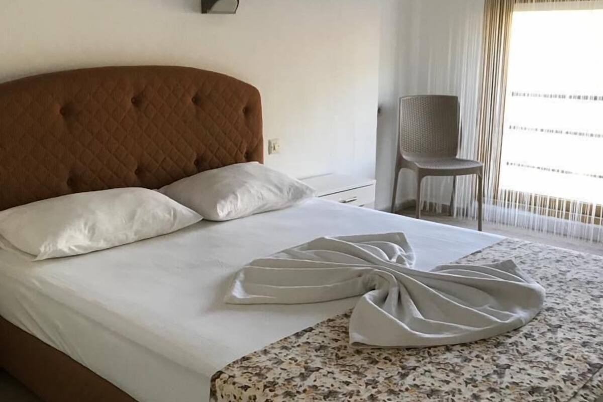 A neatly made bed with a floral bedspread and a folded white towel, a chair, and a window with blinds.