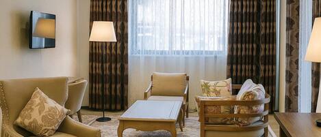 A hotel room with a sofa, armchair, and a small table. There is a window with curtains and a lamp on the wall.