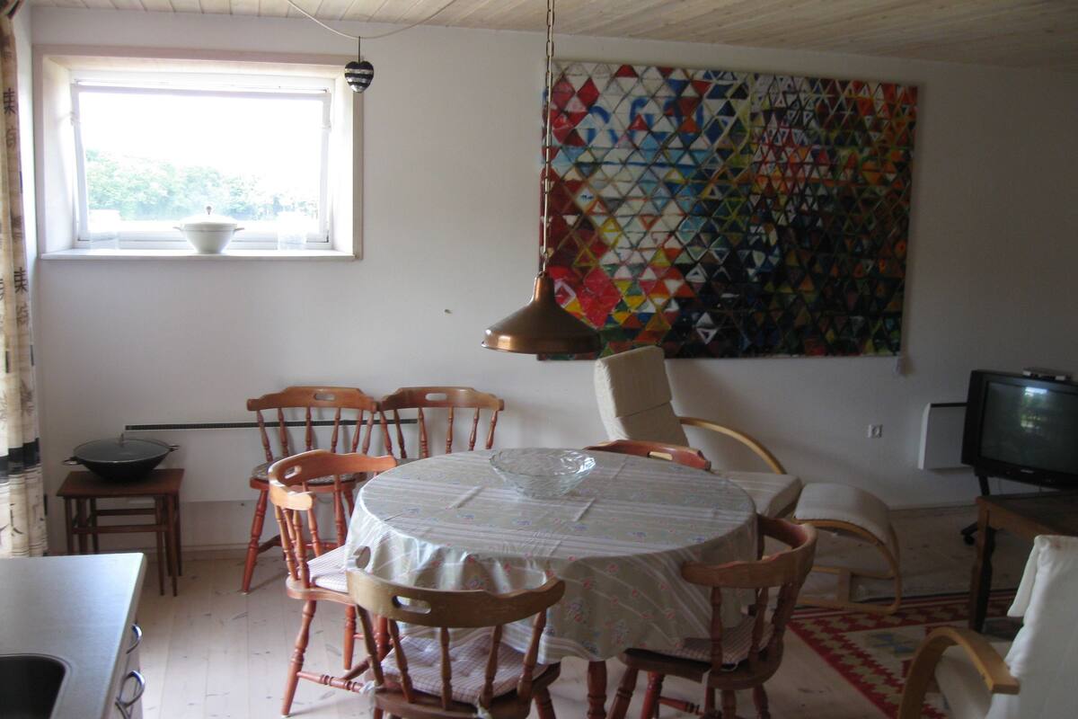 A dining area with a round table, wooden chairs, a window with a view, a colorful quilt on the wall, and a television.