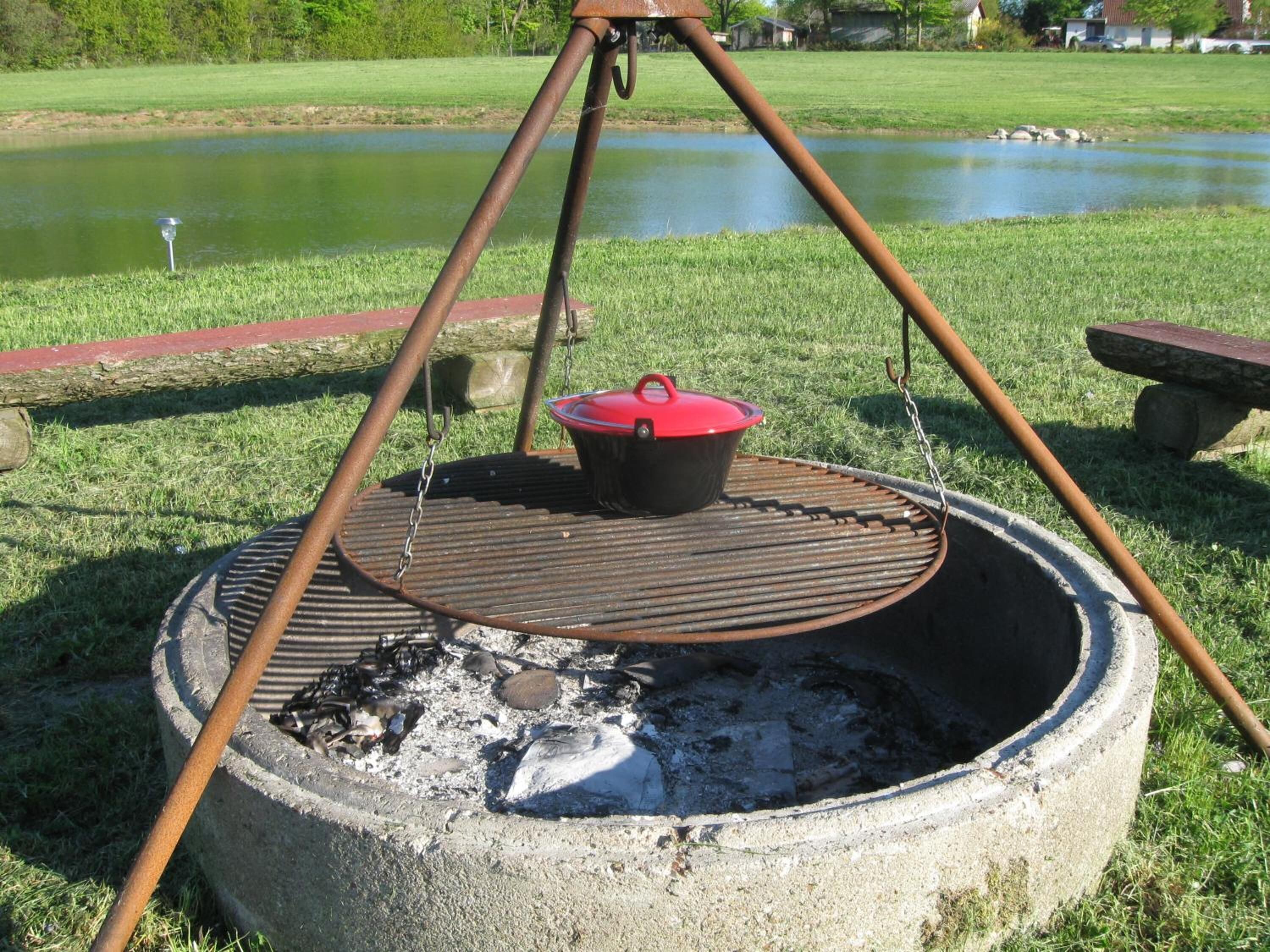 A portable outdoor grill with a pot on top, set up on a grassy area near a body of water.