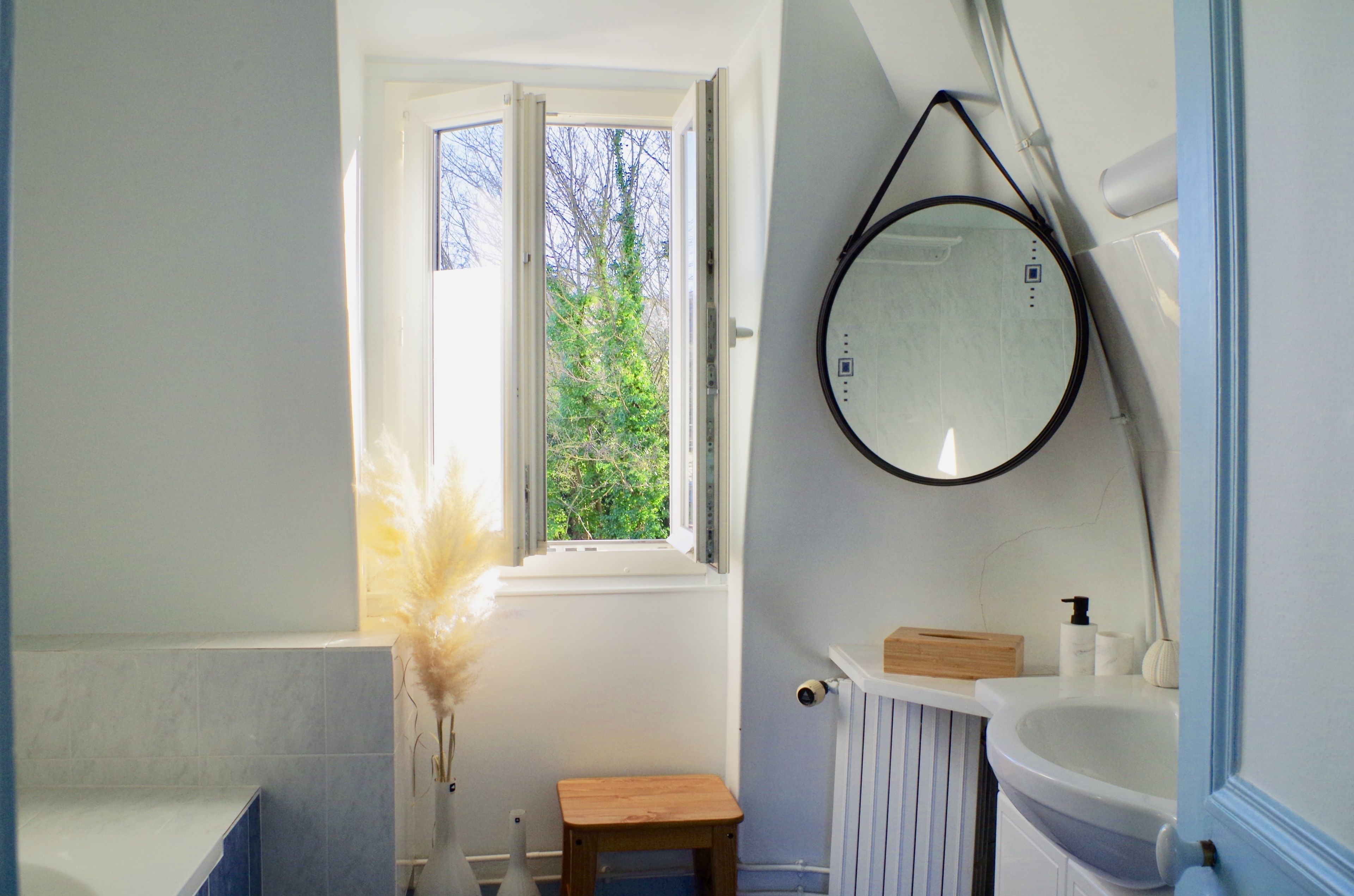 Un baño con ventana, espejo redondo, lavamanos y un taburete de madera pequeño.