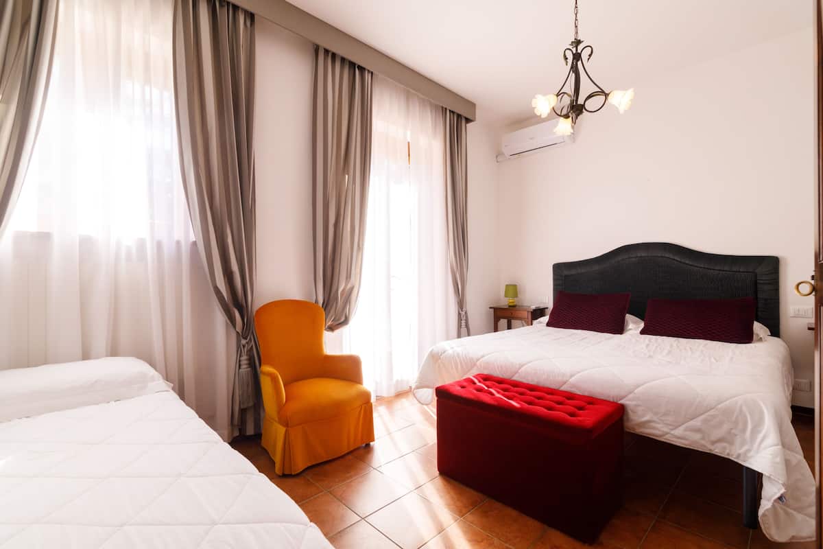 A hotel room with two beds, a red ottoman, a chair, and a chandelier.