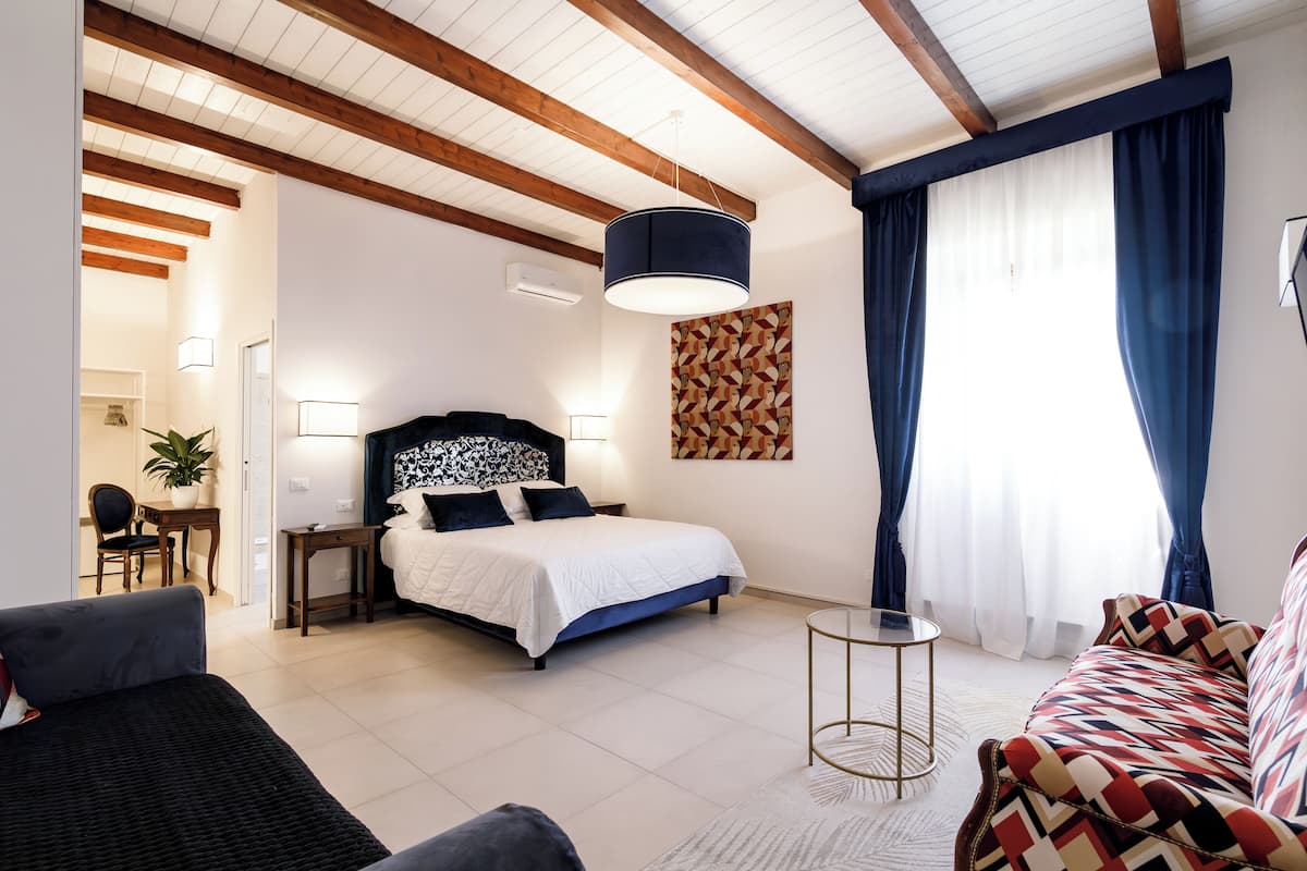 A modern bedroom with a bed, a sofa, a small table, and a desk. There is a window with curtains and a painting on the wall.