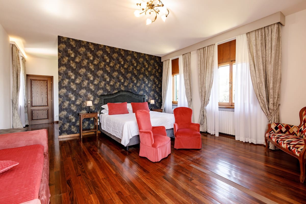A bedroom with a large bed, two red chairs, a wooden floor, and a patterned wall.