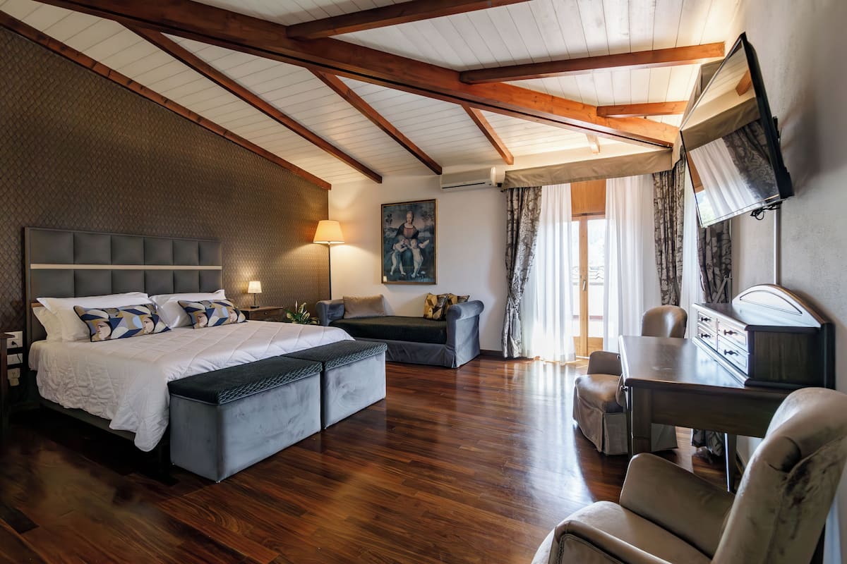 A spacious bedroom with a large bed, a desk, and a chair. There is a painting on the wall and a window with curtains.