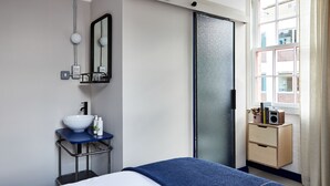A modern hotel room with a bed, a bedside table with a white bowl, a mirror, a sliding door, a window with a view of buildings, and a small wooden nightstand with a speaker. - The Buxton (London)