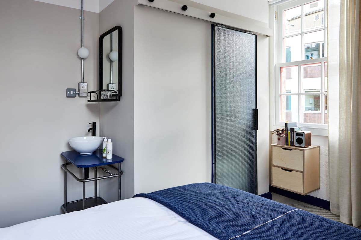 A modern hotel room with a bed, a bedside table with a white bowl, a mirror, a sliding door, a window with a view of buildings, and a small wooden nightstand with a speaker.