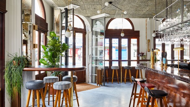 A bar area with wooden stools, a bar counter, and hanging glasses. - The Buxton (London)