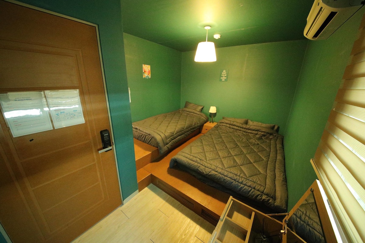 A room with two beds, a wooden door, and a small table with a lamp.