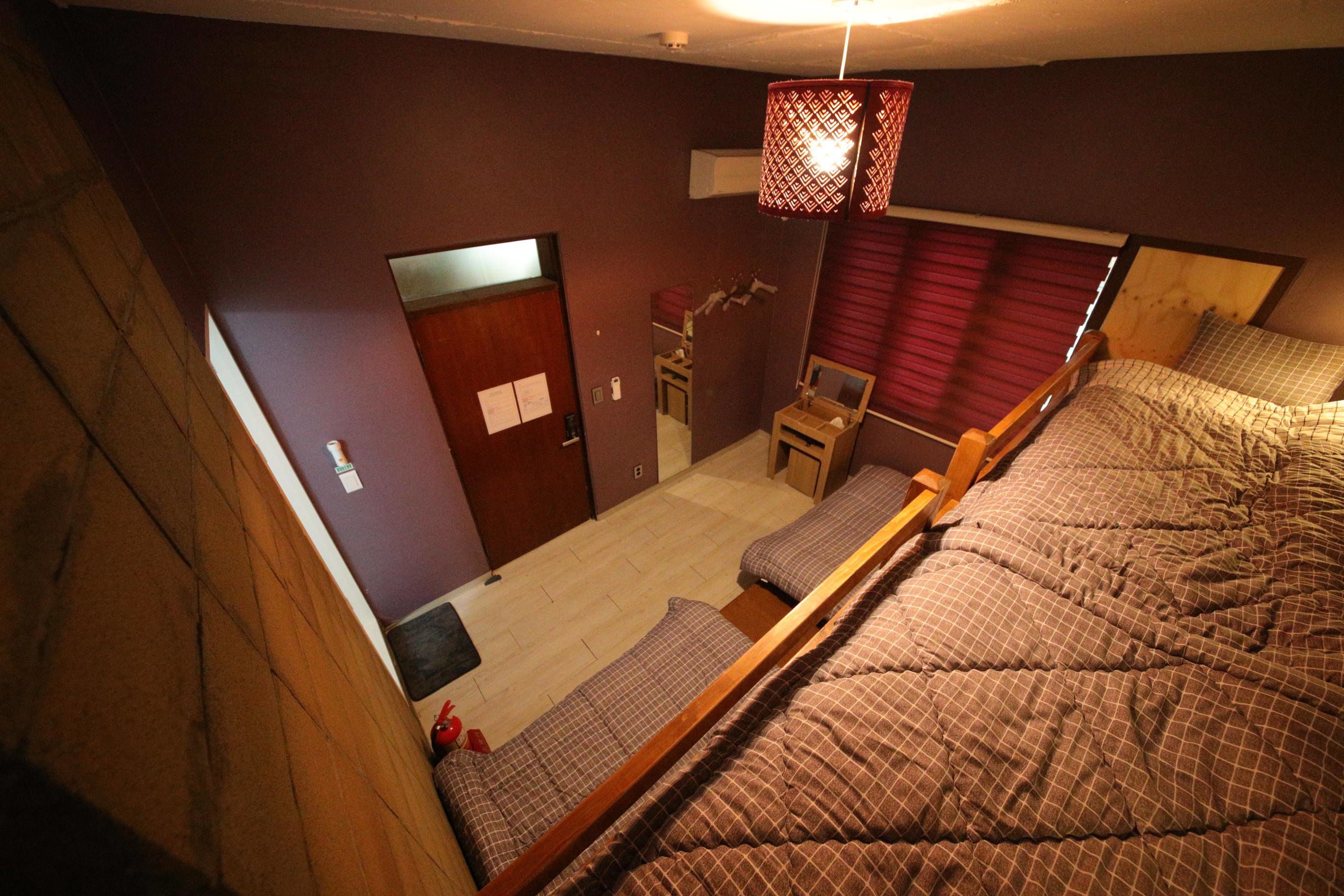 A bedroom with a bed, a desk, and a chair. There is a door and a window with curtains.