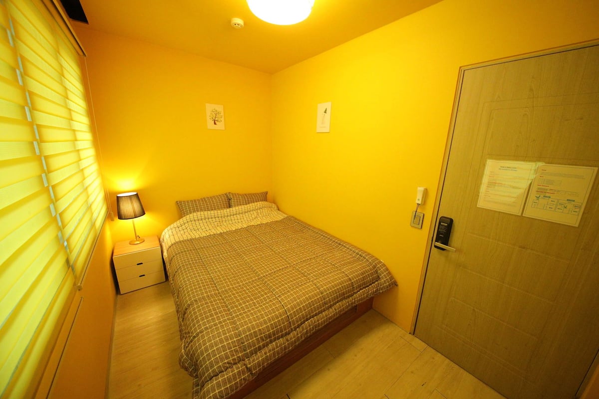 A bedroom with a bed, bedside table, lamp, and a door with a lock and papers.