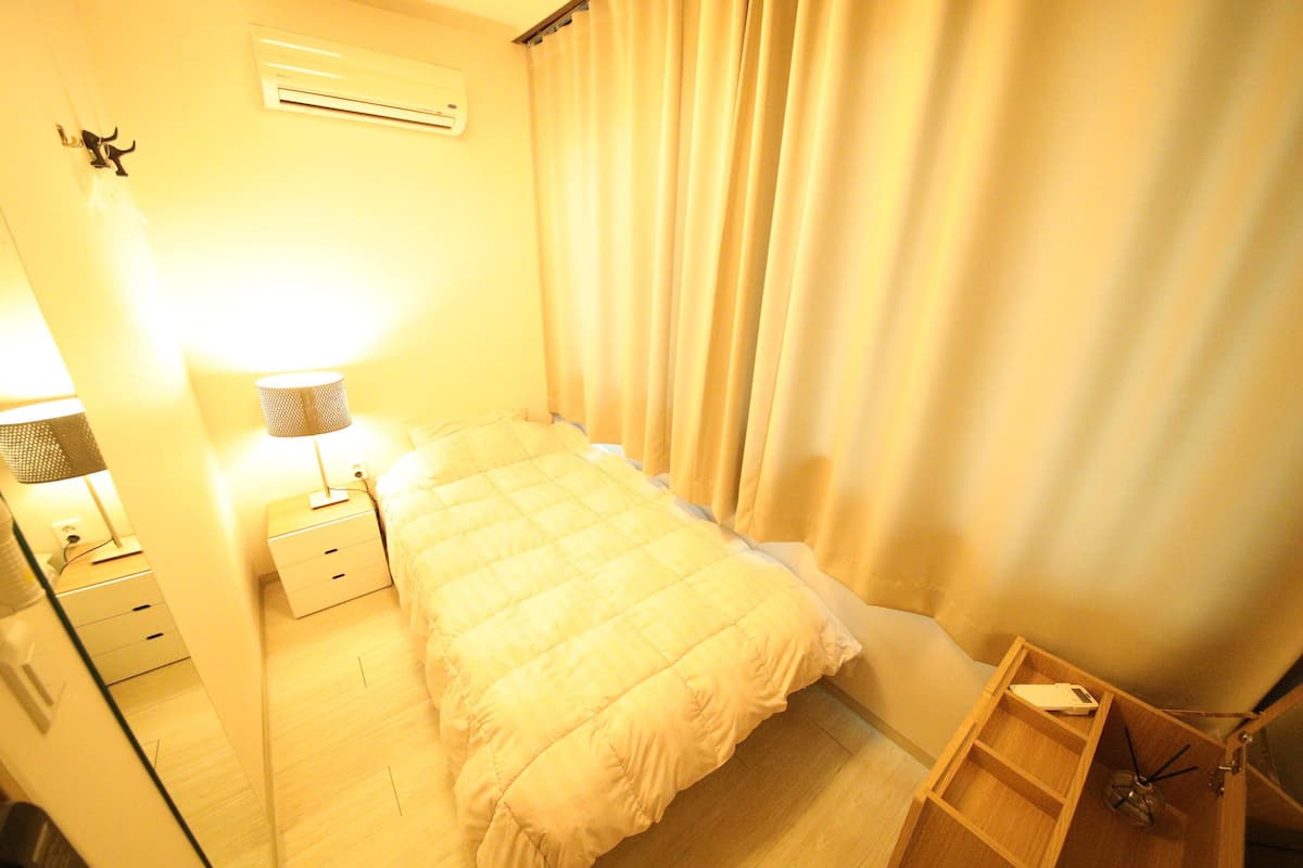 A small, neatly arranged bedroom with a single bed, a bedside table with a lamp, a wooden nightstand, and a small wooden shelf.
