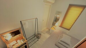 Quarto compacto com uma cama, uma escrivaninha com espelho, uma mesa de cabeceira pequena e uma janela com persianas.