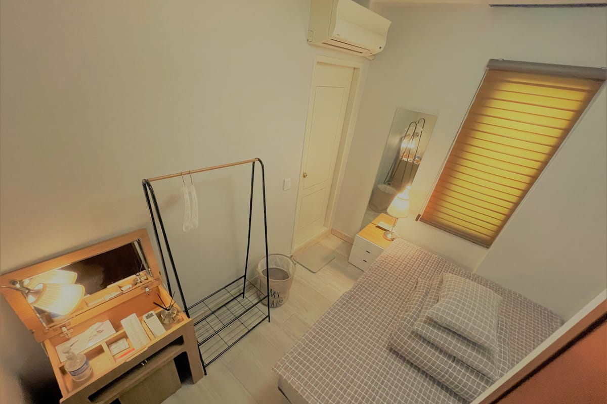 A compact bedroom with a bed, a desk with a mirror, a small bedside table, and a window with blinds.