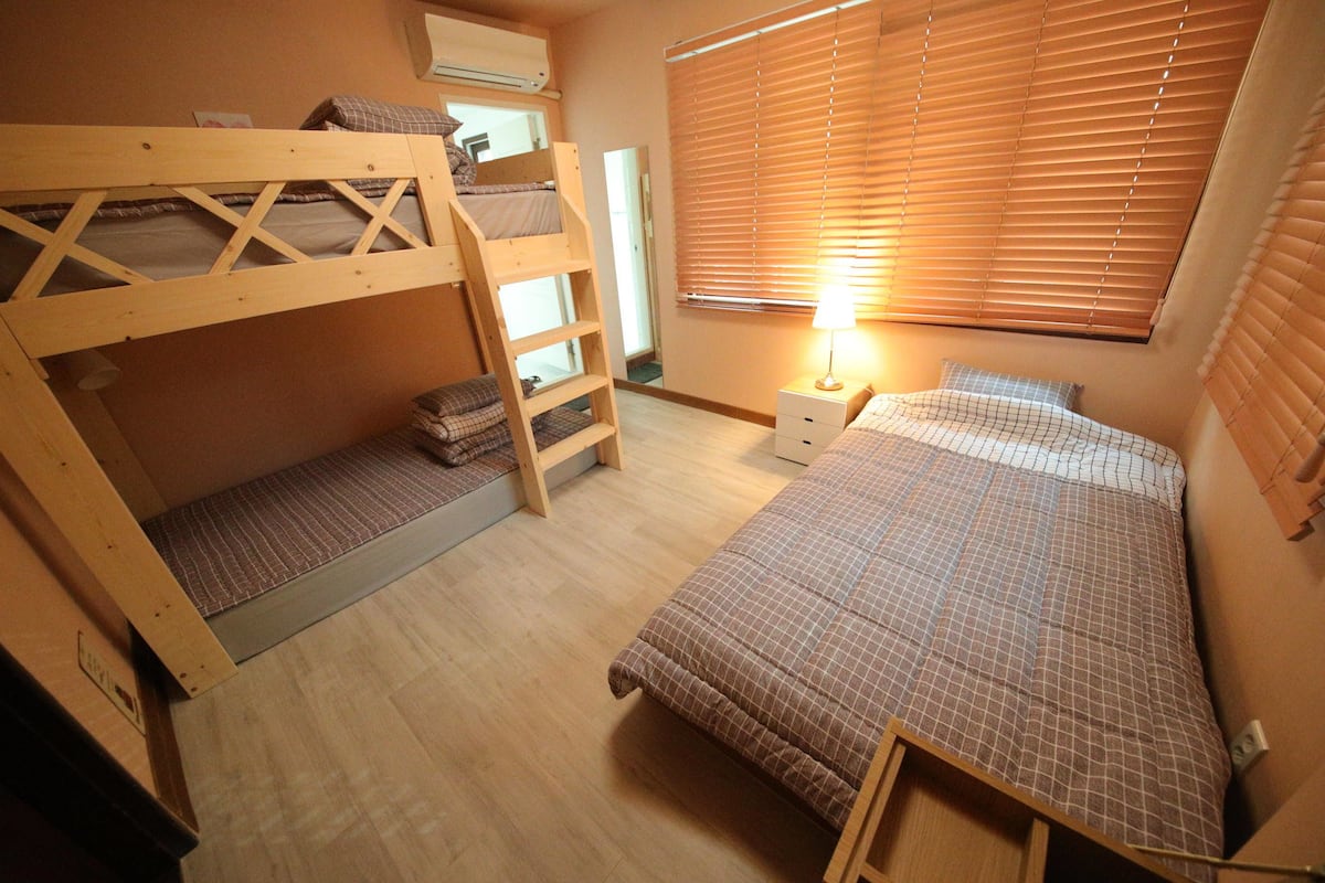 A bunk bed room with a ladder, a bedside table with a lamp, and a window with blinds.