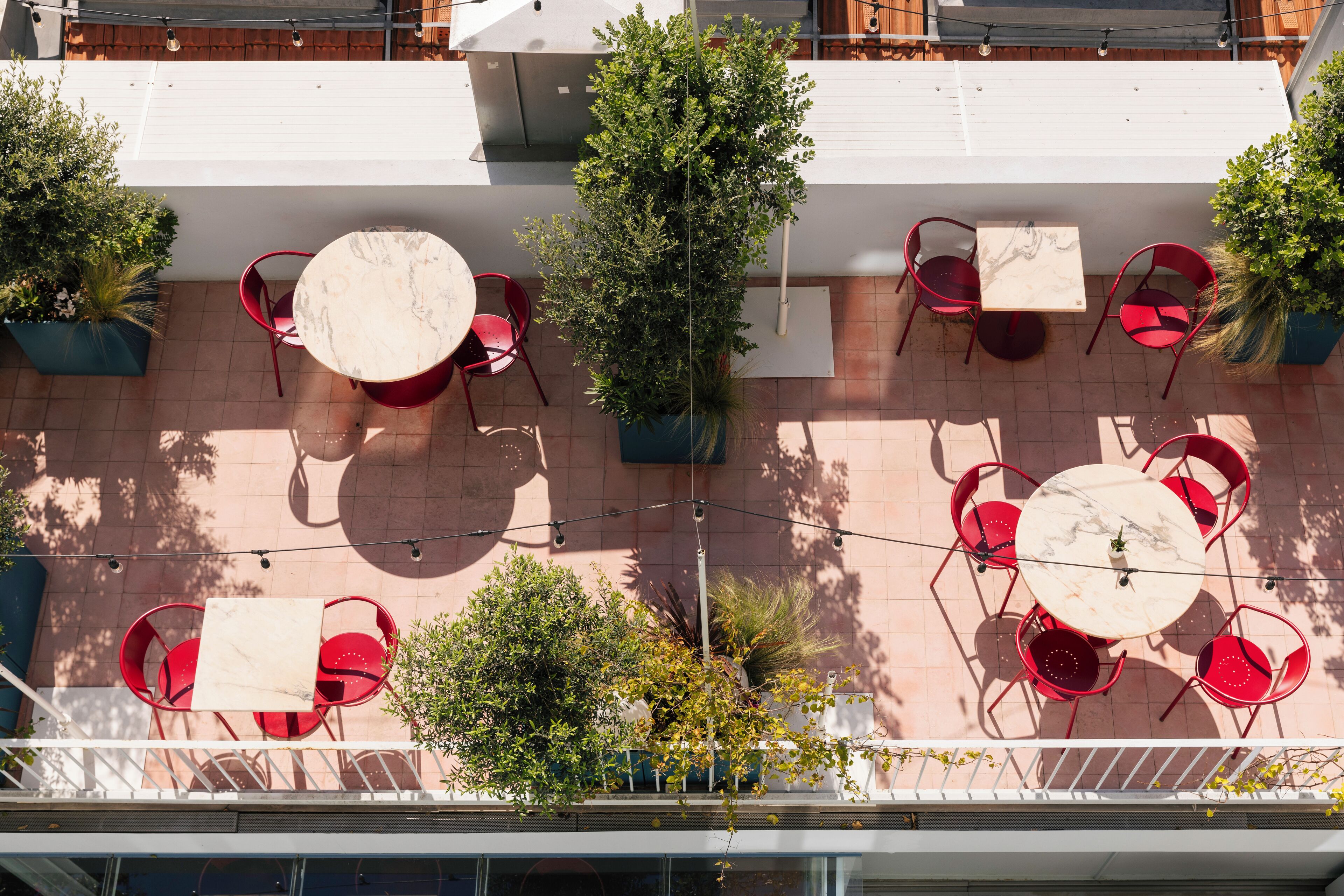 An outdoor patio with red chairs and tables, surrounded by potted plants and a white railing.