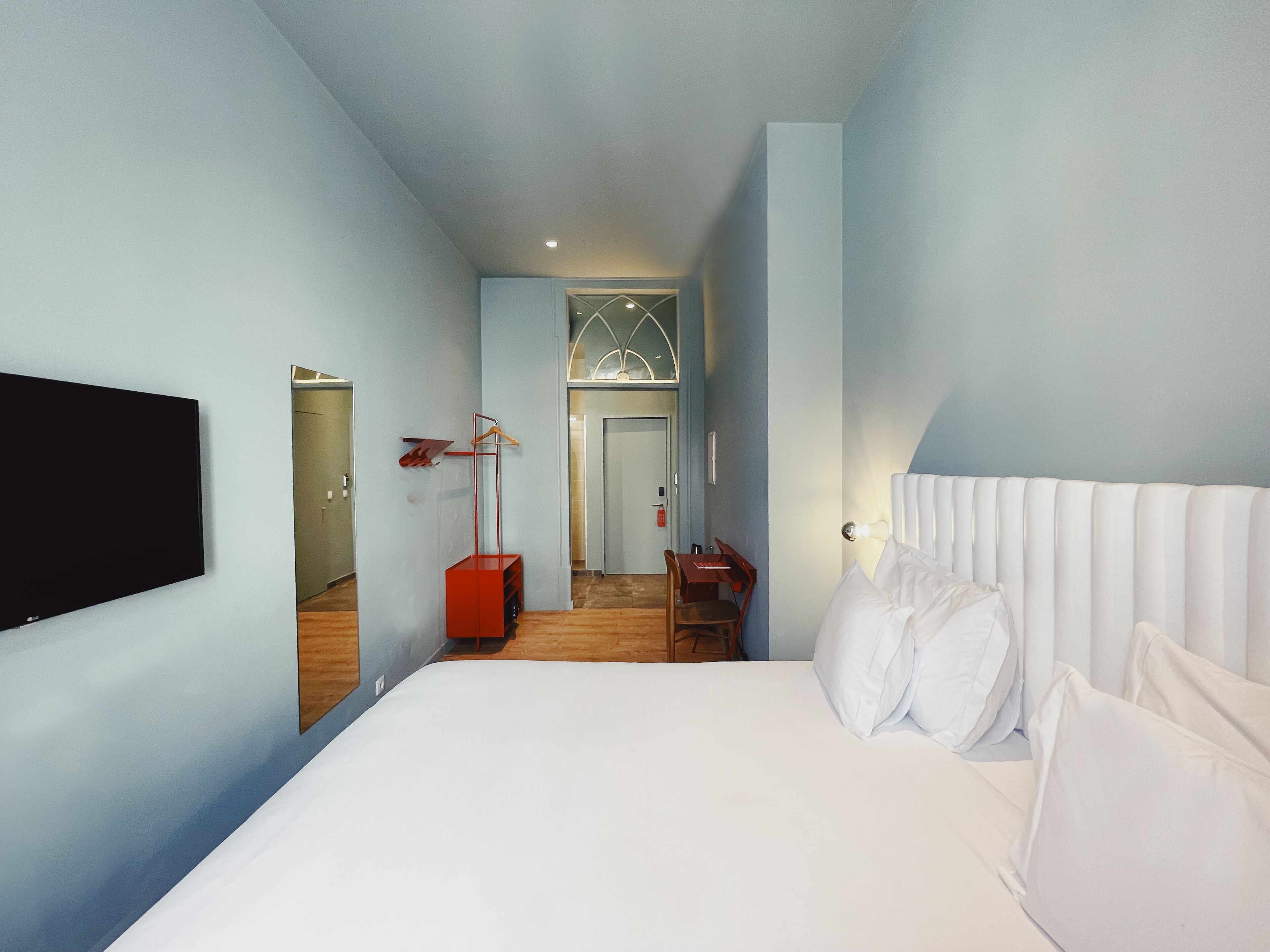 A modern hotel room with a large bed, a flat-screen TV mounted on the wall, a desk with a chair, and a mirror.