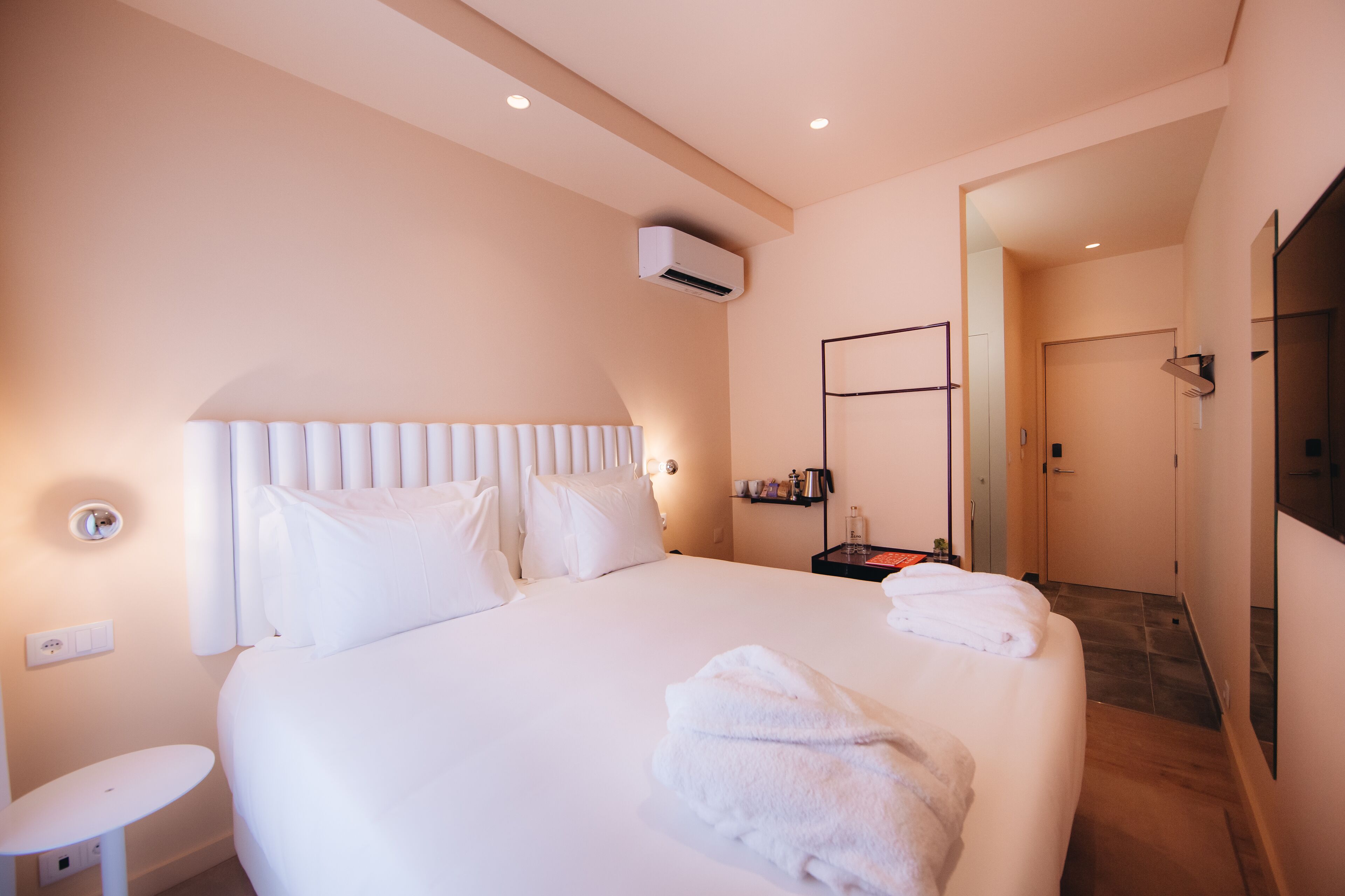 A hotel room with a large bed, bedside table, and a small table. There is an air conditioning unit on the wall and a door leading to another room.