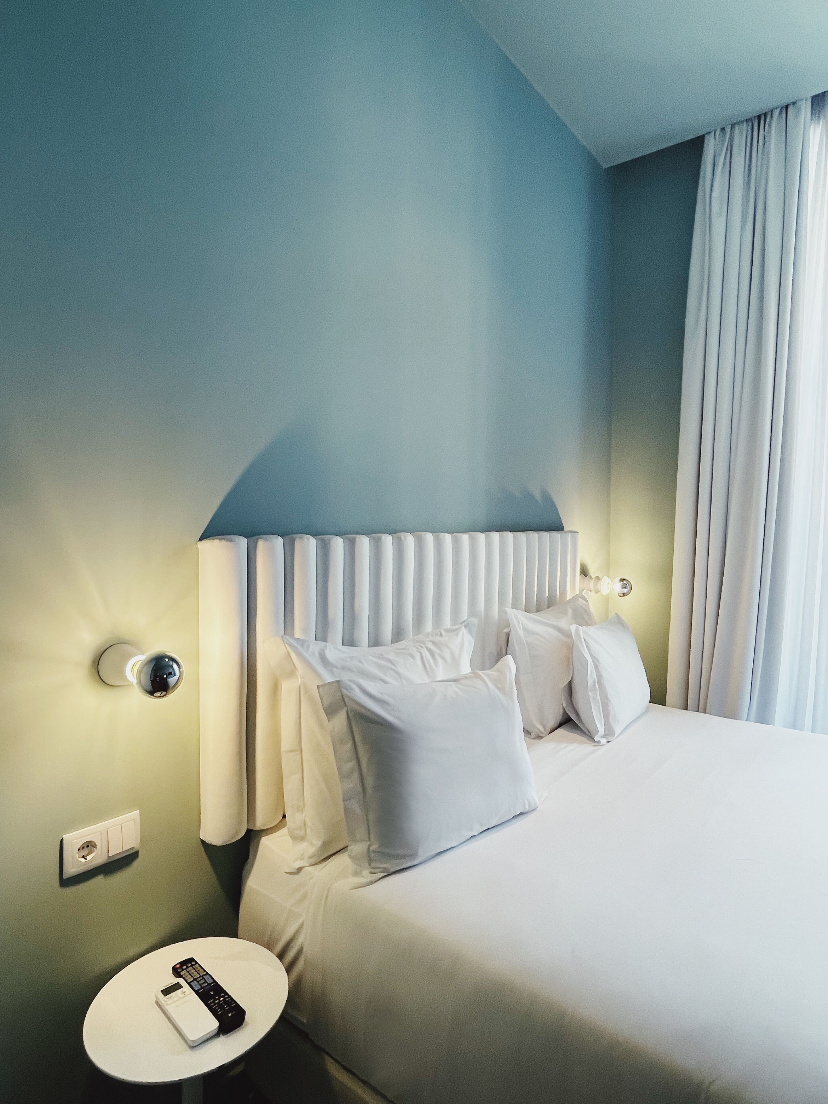 A hotel room with a bed, white bedding, a round bedside table with a remote control, and a wall-mounted light fixture.