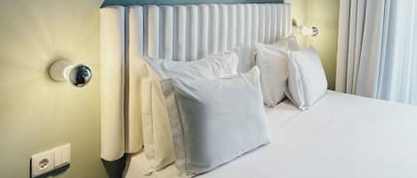 A hotel room with a bed, white bedding, a round bedside table with a remote control, and a wall-mounted light fixture.