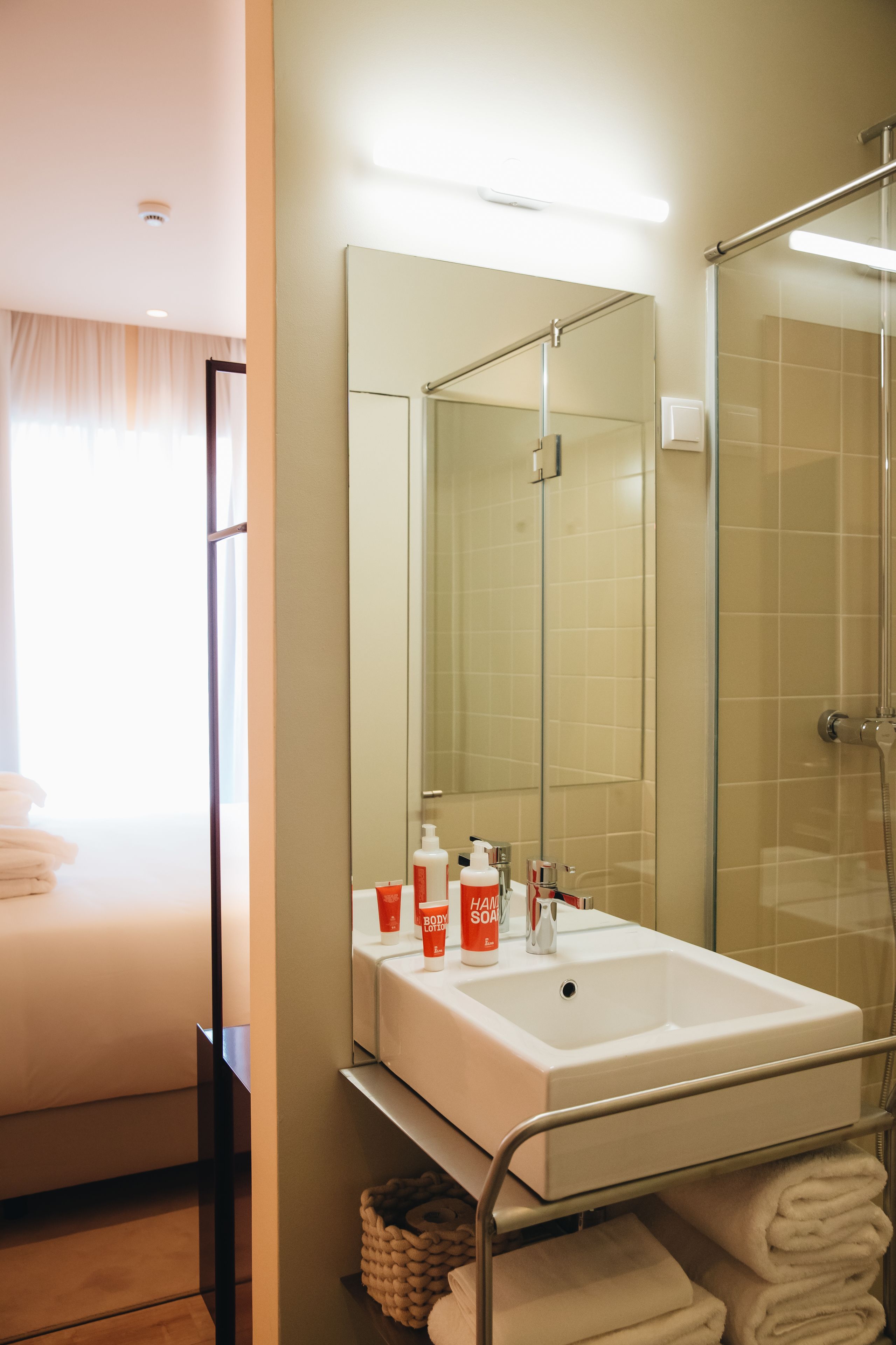 A bathroom with a white sink, a mirror, a glass shower enclosure, and a bed with white linens.