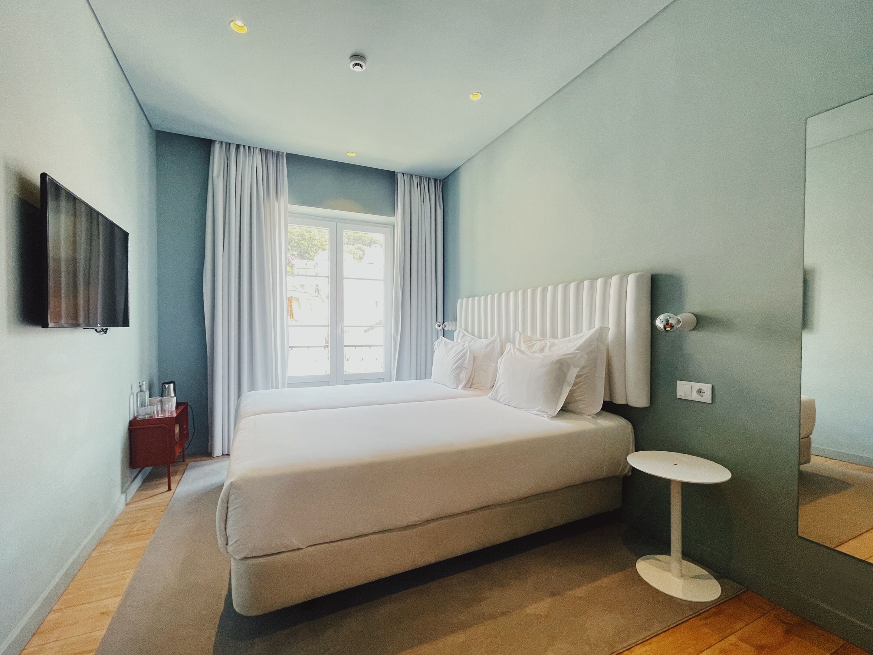 A modern bedroom with a large bed, a bedside table, a TV mounted on the wall, and a window with curtains.